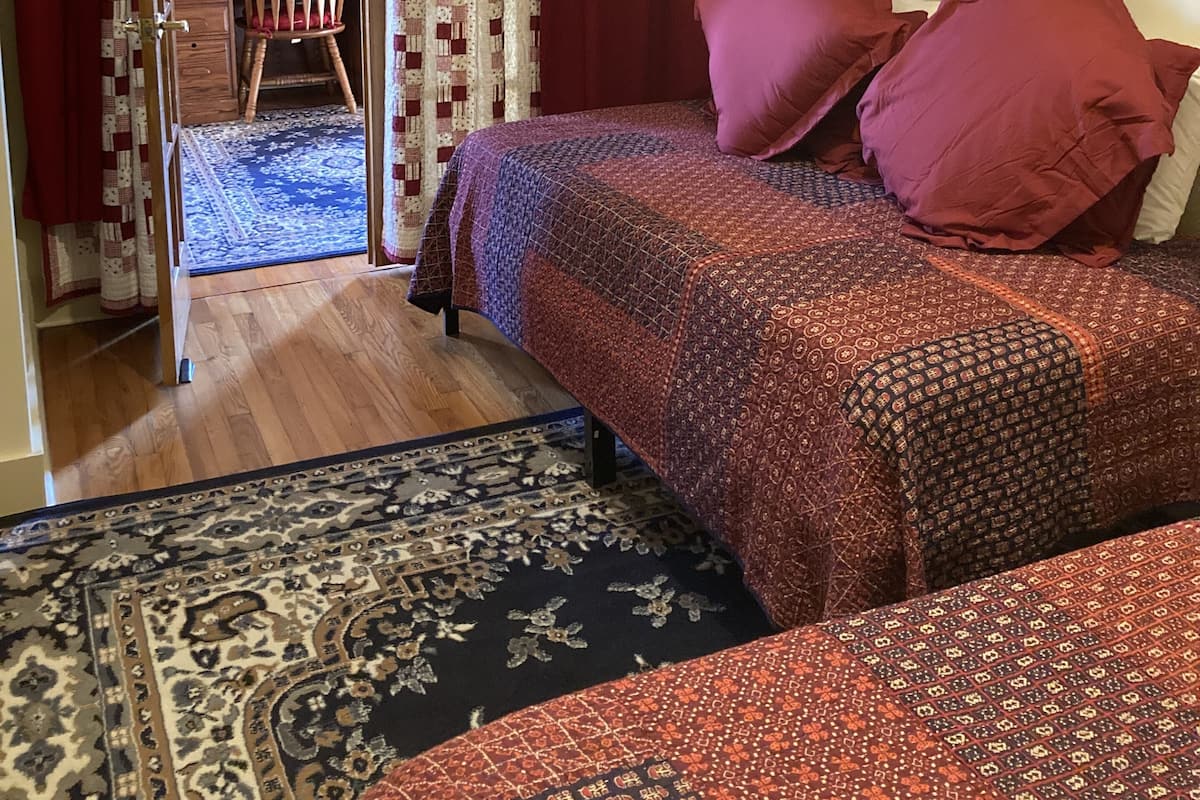 Quarto com cama, duas almofadas, uma cadeira, escrivaninha e tapete estampado.