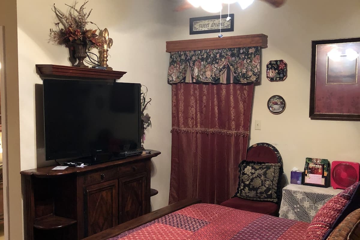 Quarto com cama, televisão sobre um móvel de madeira, ventilador de teto e cortina florida.