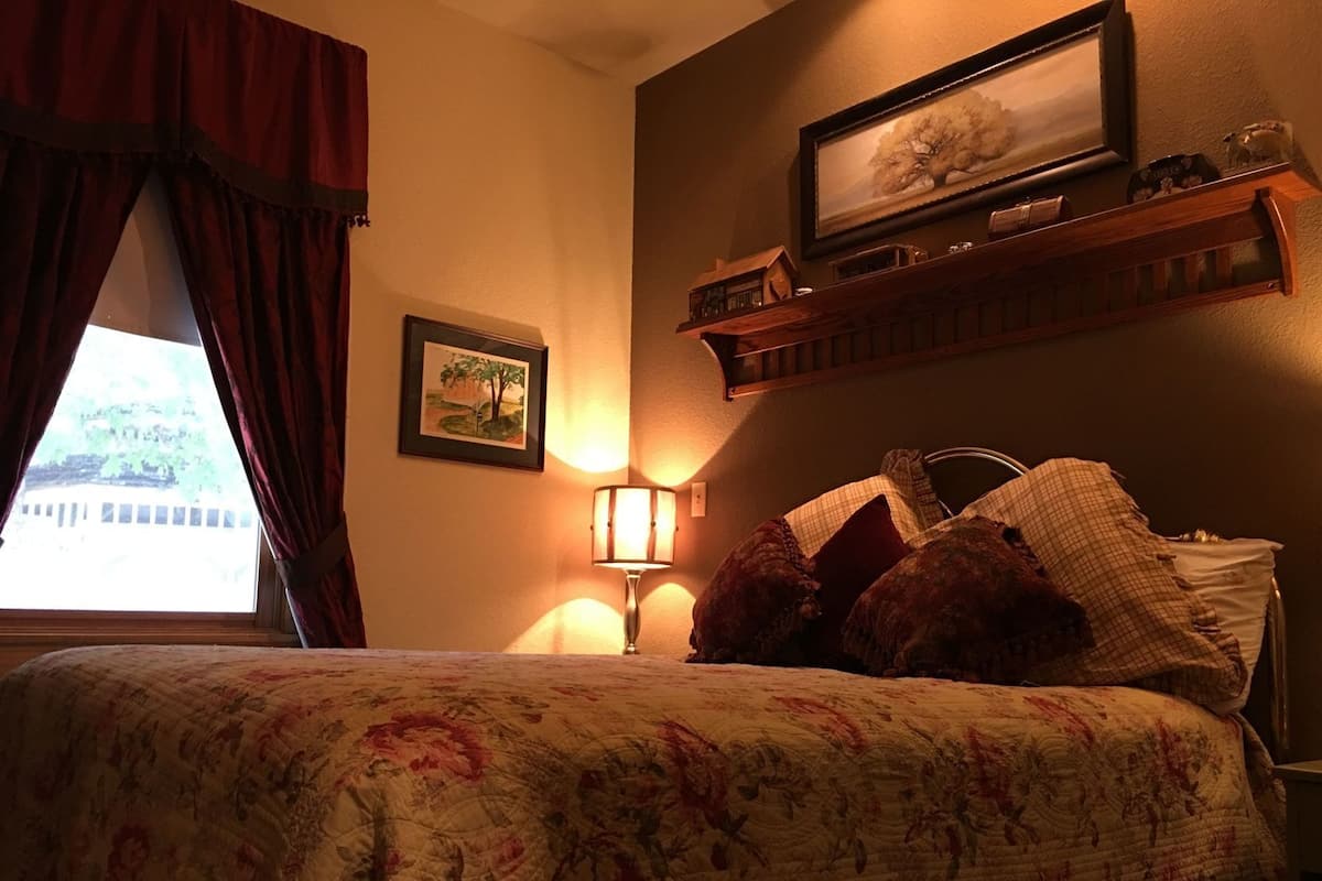Quarto de hotel com cama, luminária, quadro e janela com cortinas.
