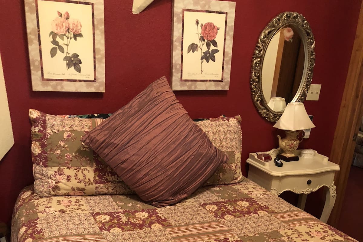 Quarto com colcha estampada com motivos florais, um travesseiro roxo, duas gravuras botânicas emolduradas na parede e um espelho sobre uma mesinha de cabeceira.