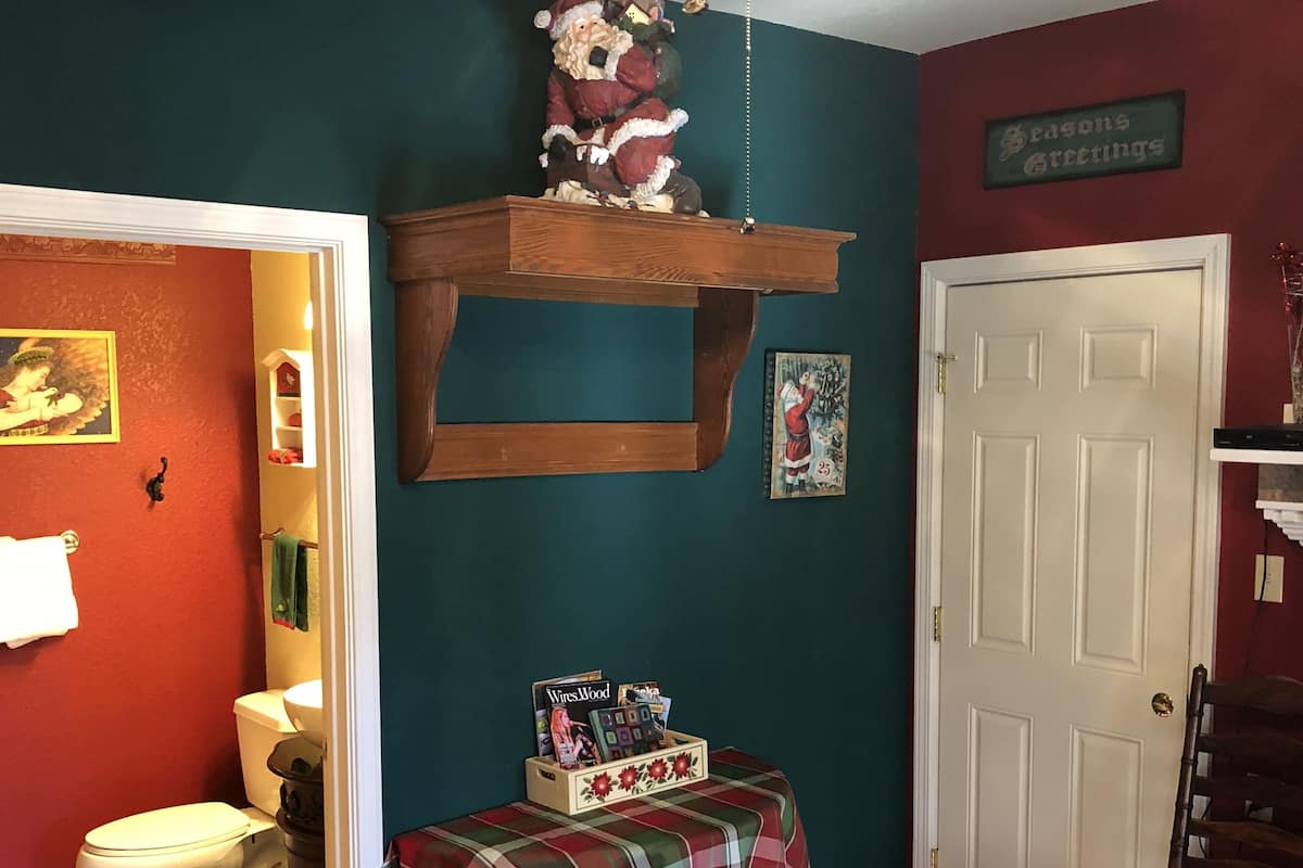 Quarto com estante de madeira que exibe um boneco do Papai Noel, mesa xadrez com revistas, porta com placa de 'Saudações Festivas' e tapete estampado.