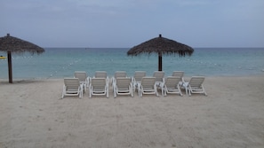 A beach with white lounge chairs arranged in rows, two thatched umbrellas, and a clear view of the sea. - Roots Bamboo Beach Resort (Negril)