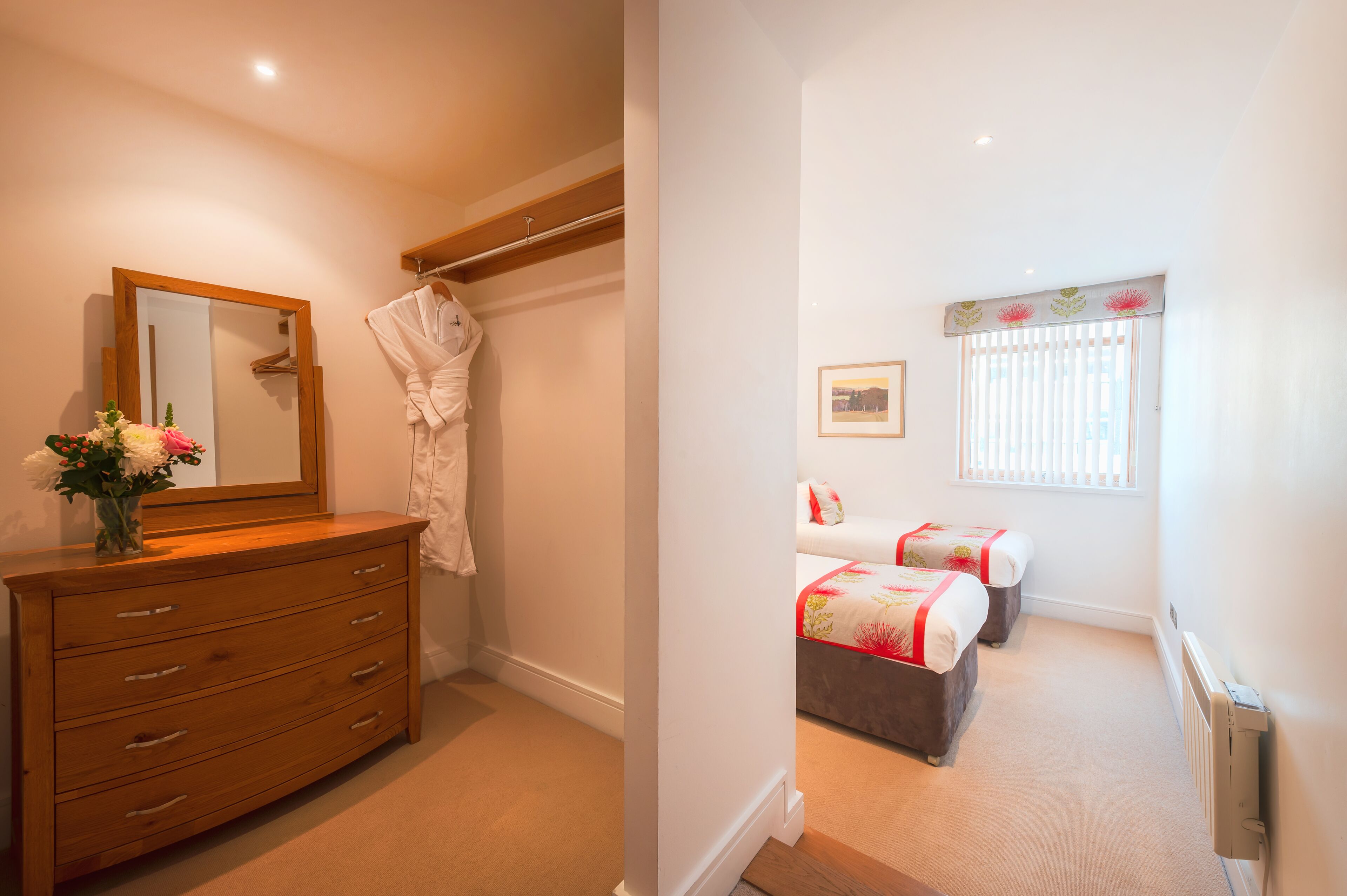 Une chambre avec une commode en bois, un miroir, un peignoir, un lit recouvert d’une couvre-lit à motifs floraux et une fenêtre avec des stores.