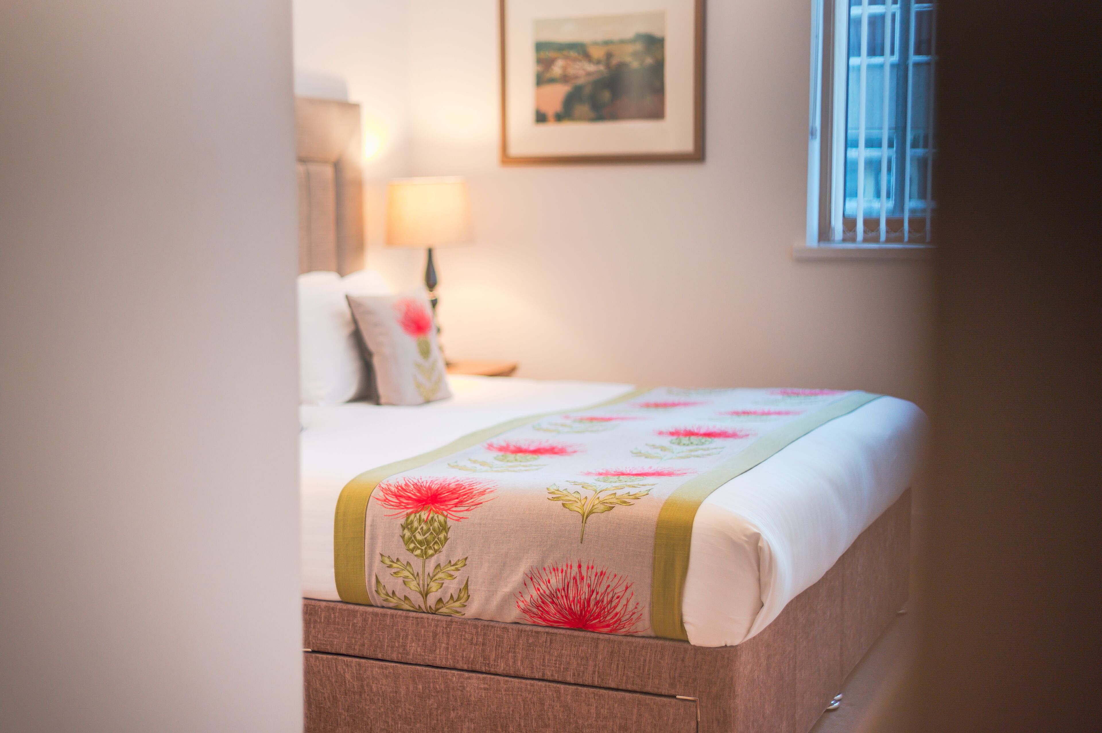 A neatly made bed with a floral bedspread, a bedside lamp, and a framed picture on the wall.