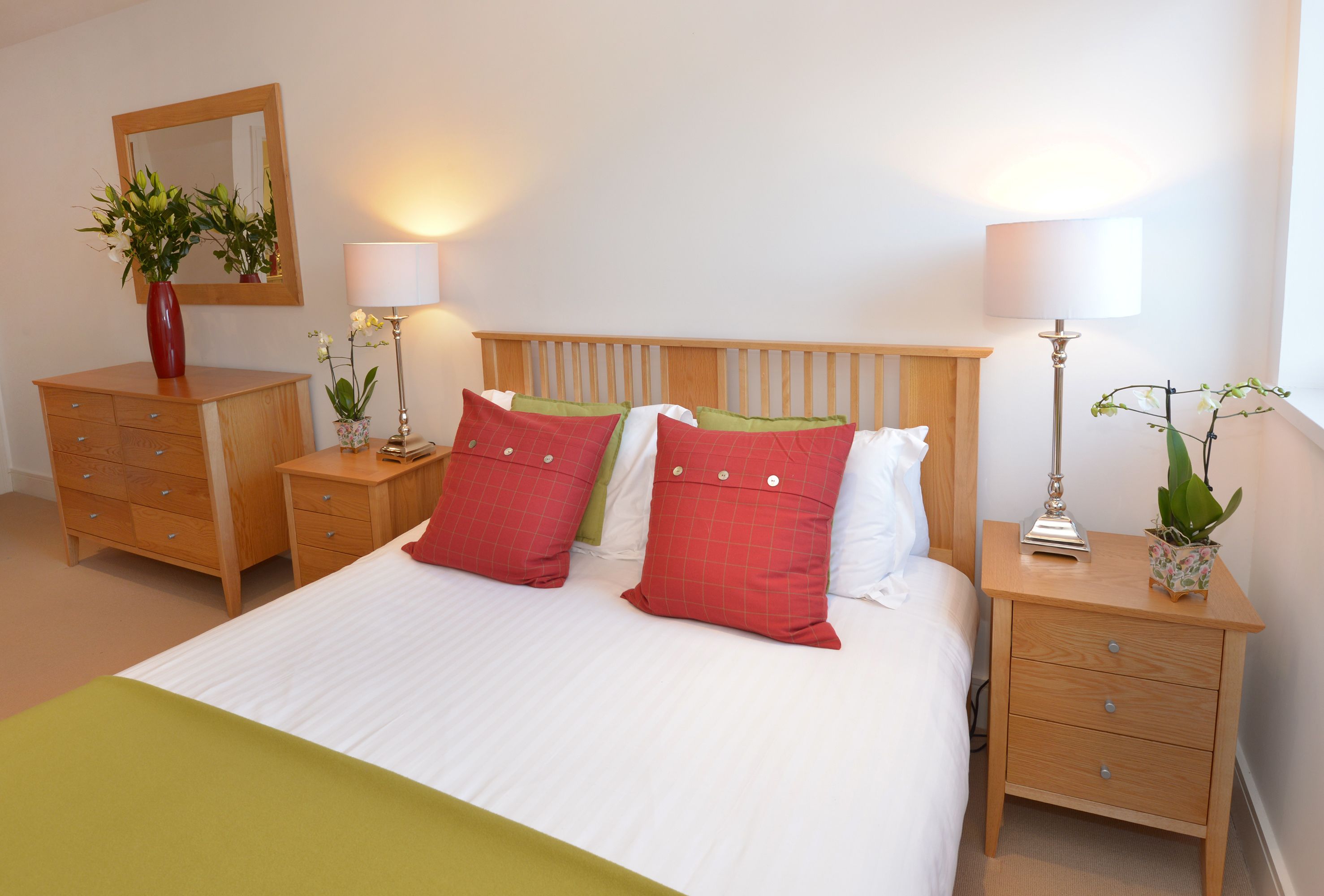 A neatly arranged bedroom with a wooden headboard, a bed with white bedding, two red pillows, a green throw, and two bedside tables with lamps and small potted plants.