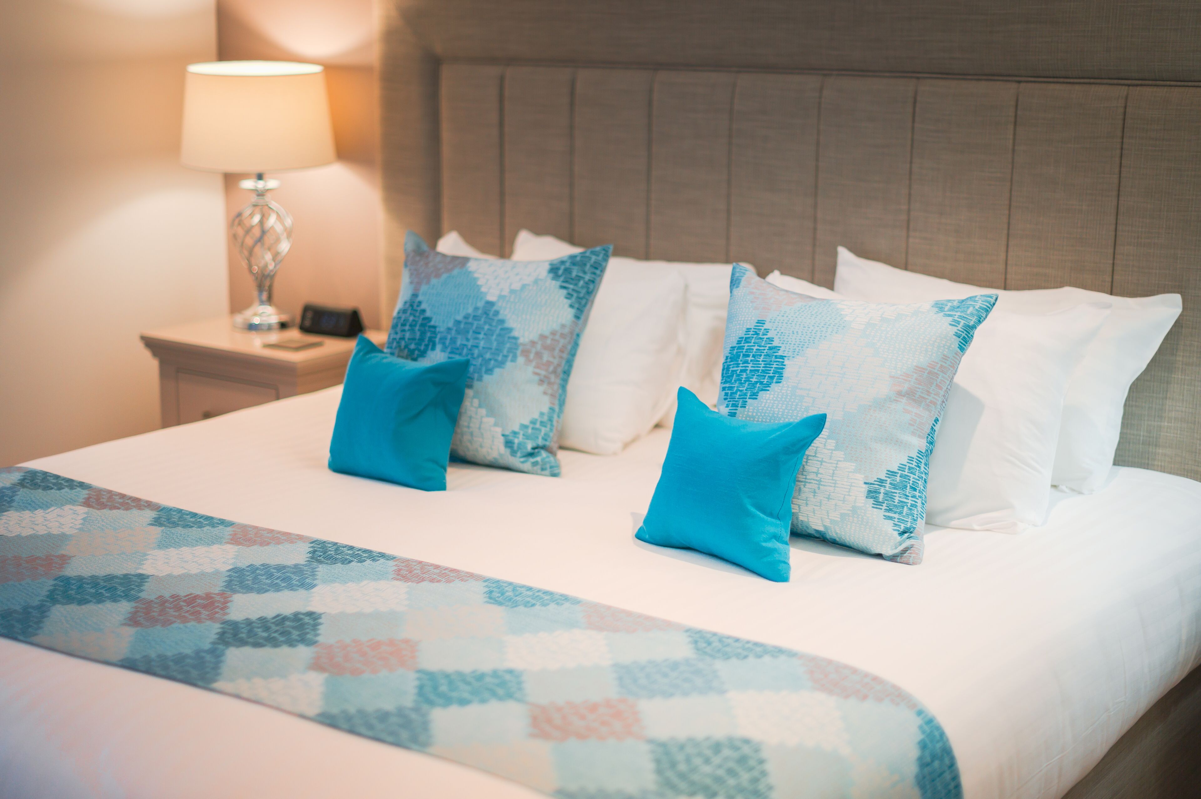 A neatly made bed with a patterned bedspread and multiple pillows, a bedside lamp, and a nightstand with a phone and a small decorative item.