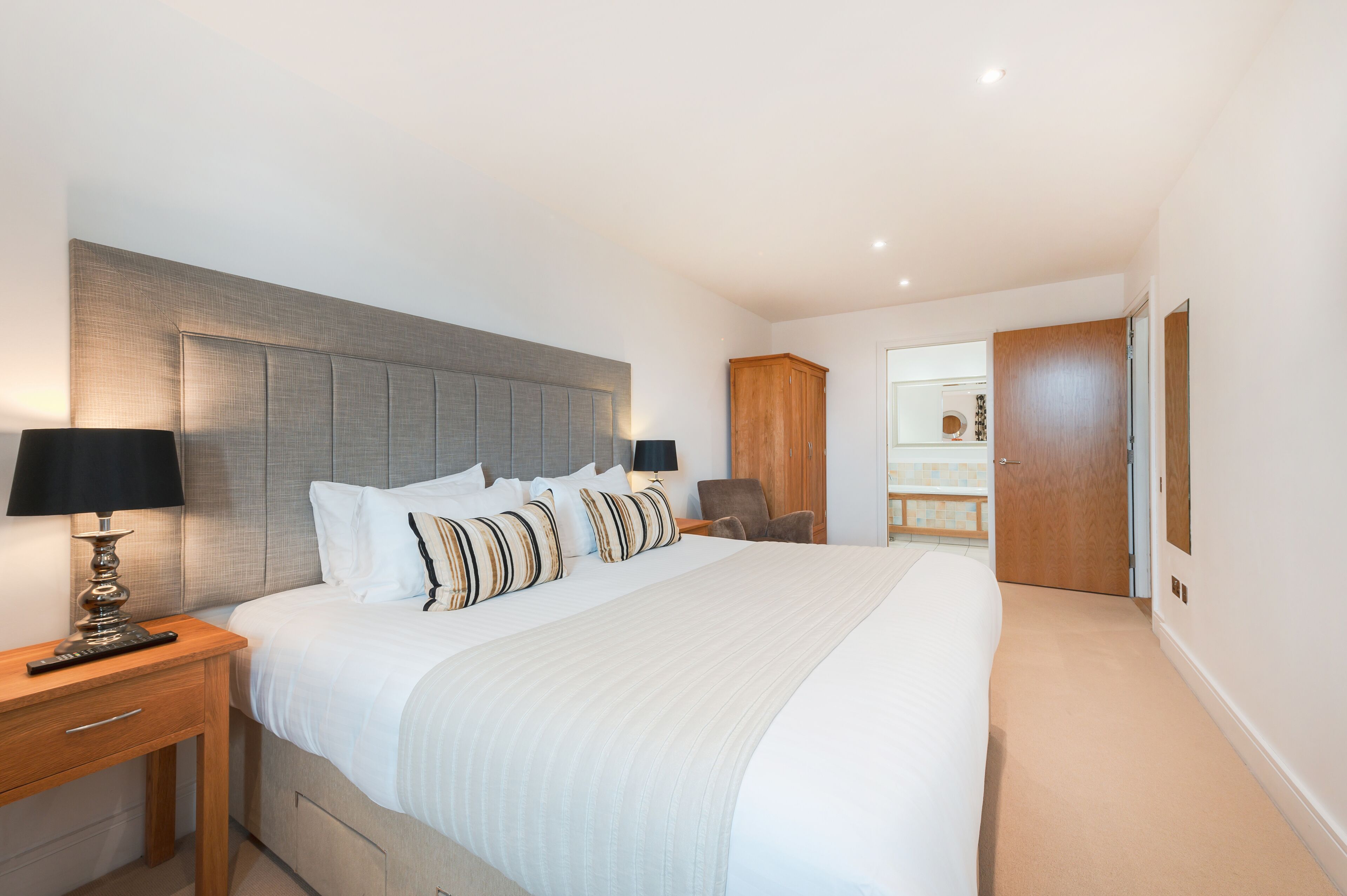 Une chambre moderne avec un grand lit, des tables de chevet, une armoire en bois et une salle de bain visible à travers une porte ouverte.