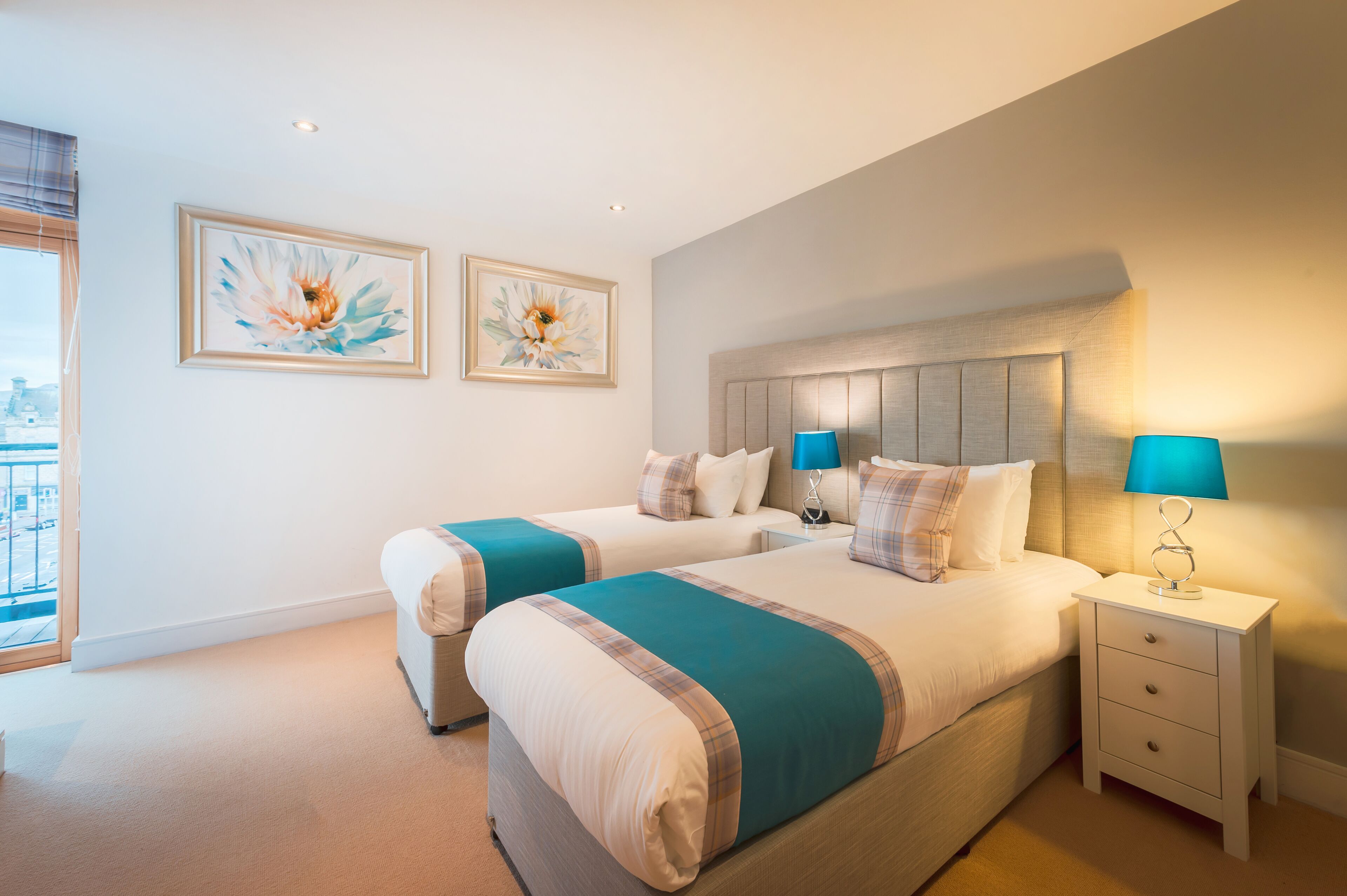 A hotel room with two beds, a nightstand with a lamp, and two framed floral paintings on the wall.