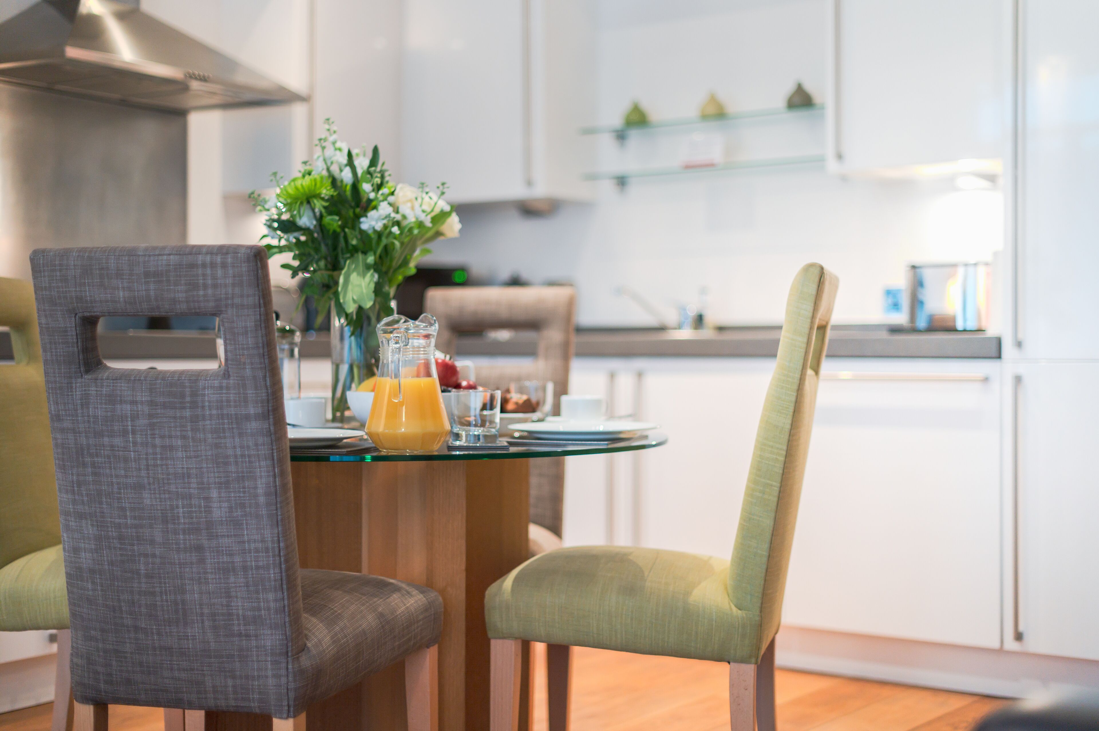 A modern kitchen with a dining table set for two, featuring a vase with flowers and a glass of orange juice.