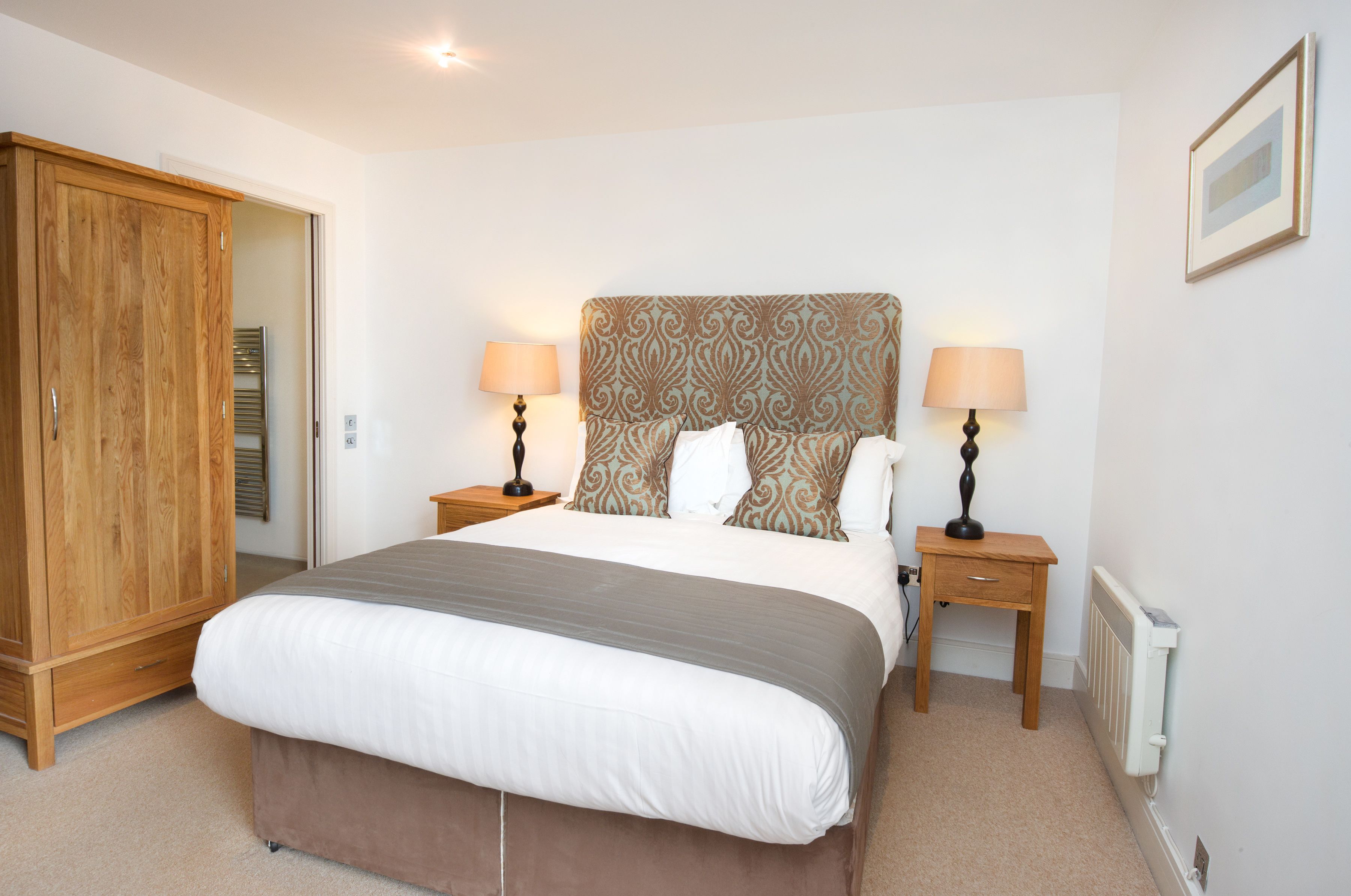 A bedroom with a large bed, two bedside tables with lamps, a wooden wardrobe, and a radiator.
