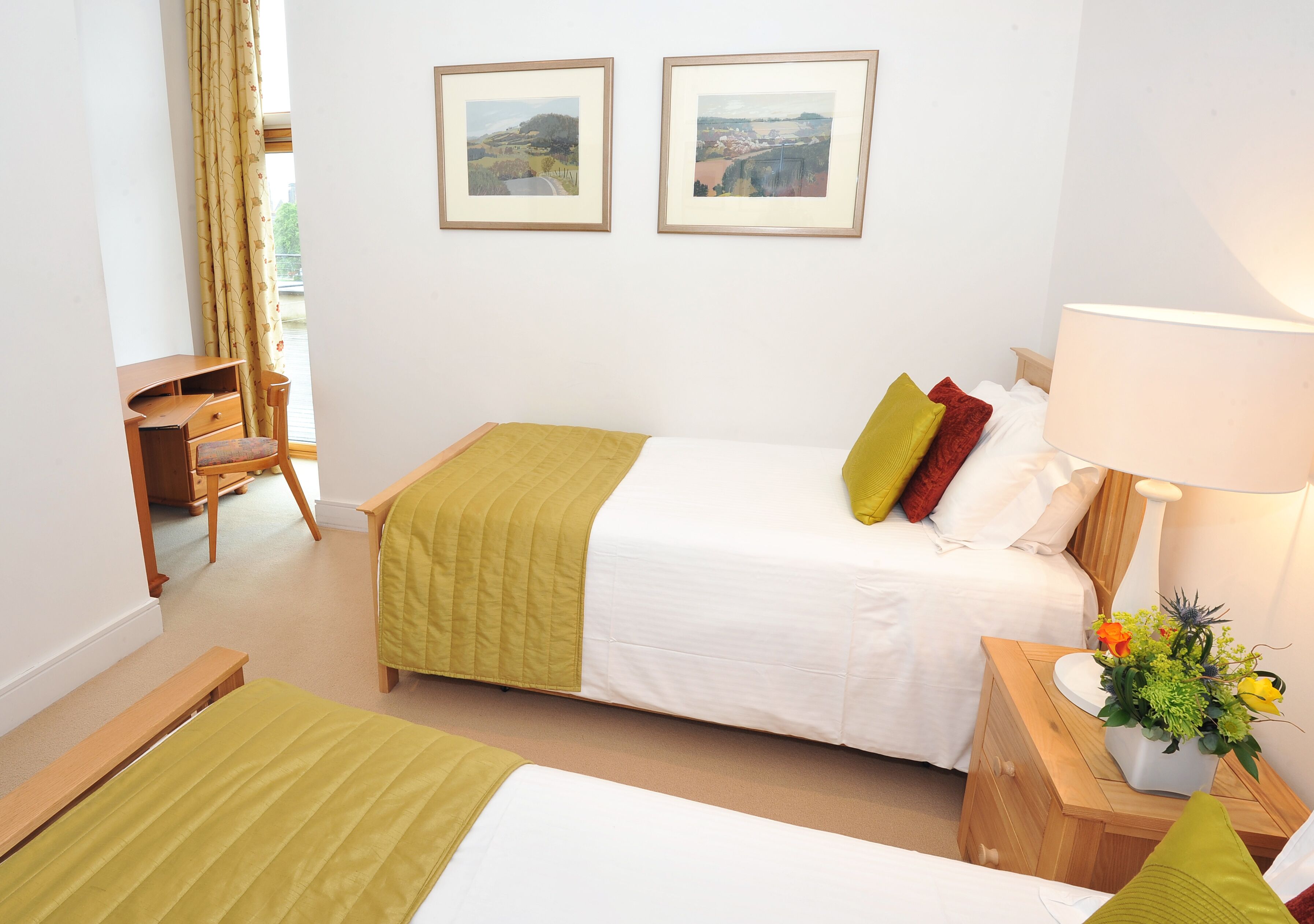 Een hotelkamer met twee bedden, een bureau, een stoel, een lamp en uitzicht op het buiten.