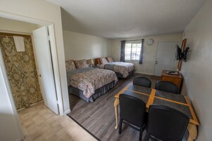 Una habitación con dos camas, una mesa de comedor con sillas, una mesita con lámpara y una puerta que da a un baño.
