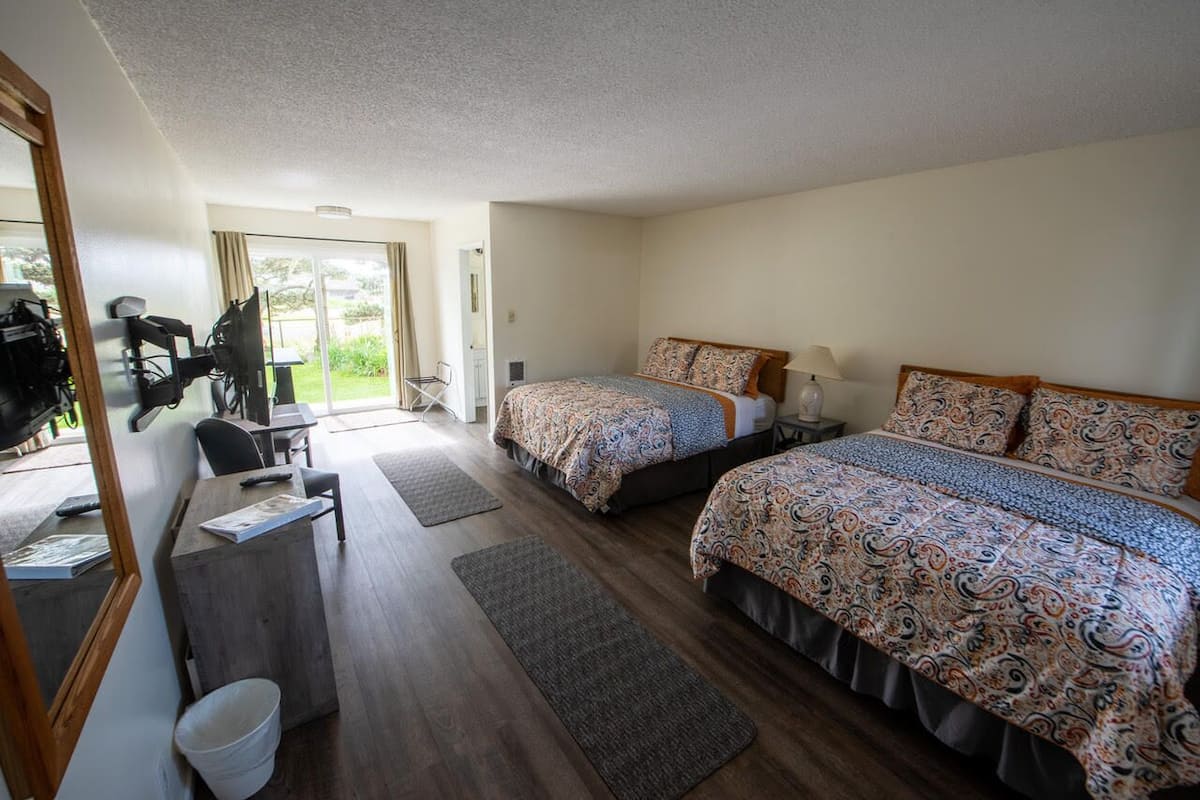 A hotel room with two beds, a desk, and a chair. There is a window with a view of greenery outside.