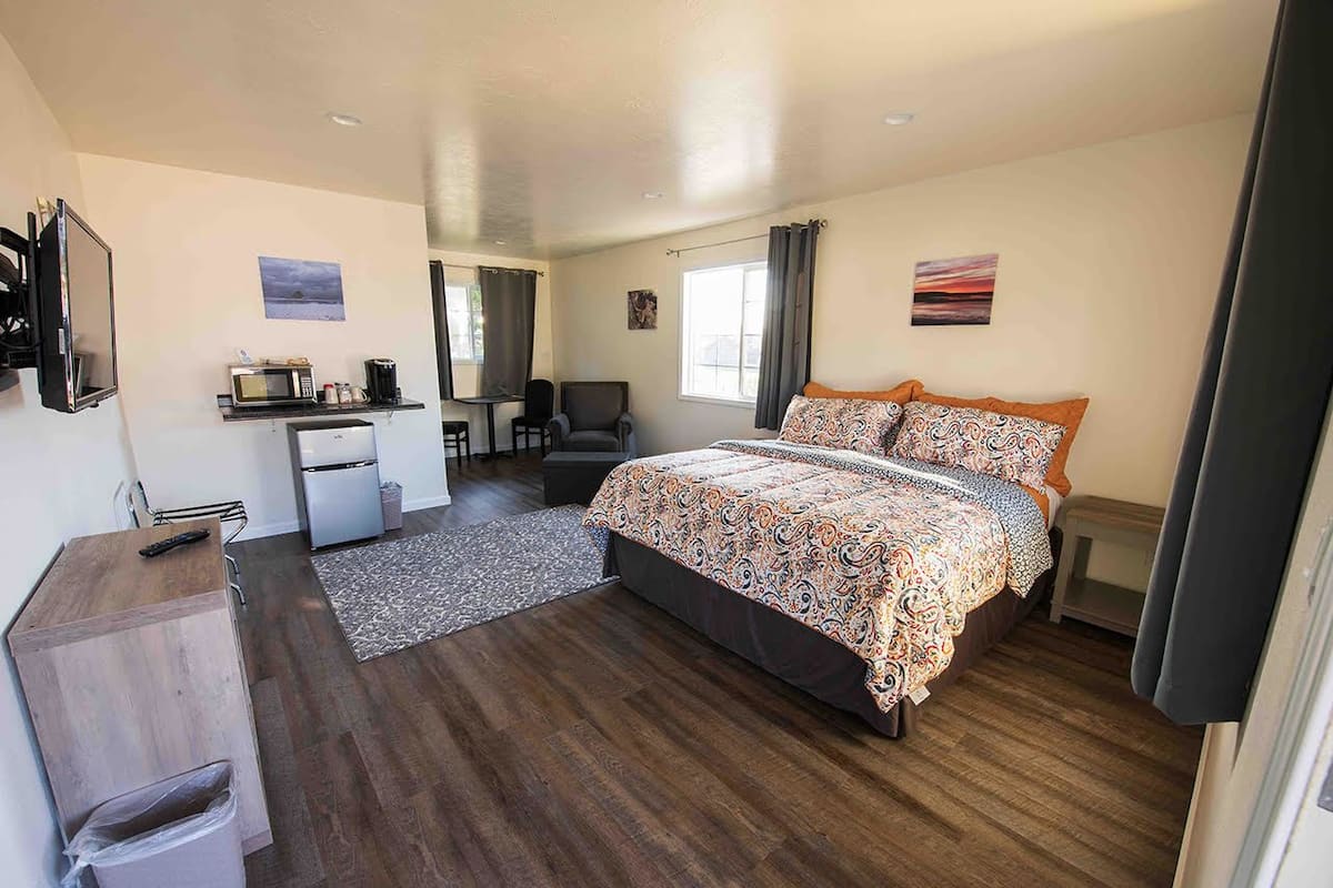 A bedroom with a bed, a desk, a chair, a small kitchenette, and a trash bin.