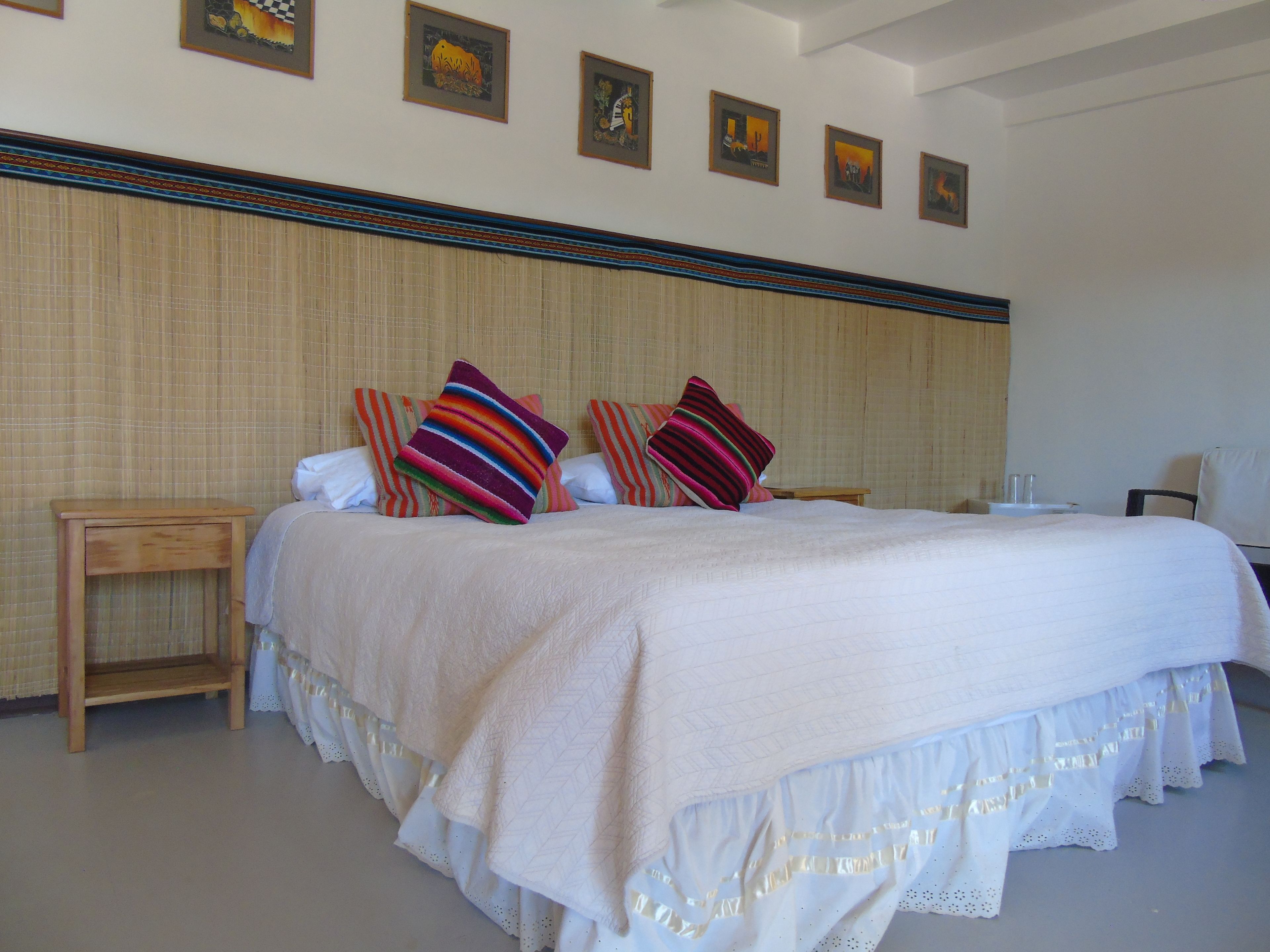 Una cama bien tendida con edredón blanco y varios cojines de colores, una mesita de noche de madera y una pared decorada con cuadros.