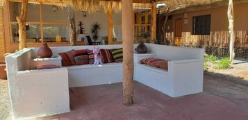 Un sofá blanco con cojines coloridos, un jarrón grande y un peluche en un espacio exterior iluminado por el sol, con techo de paja y pilares de madera.