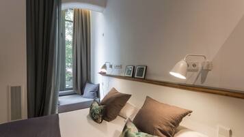 Un dormitorio con cama, almohadas, mesita de noche con lámpara, ventana con cortinas y estante en la pared con fotografías.