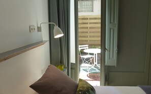 A room with a bed, a bedside lamp, a window with curtains, and a view of an outdoor patio with a table and chairs. - Casa Mathilda (Barcelona)