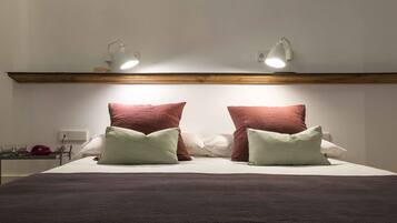 A neatly made bed with two large pillows and two smaller pillows, a bedside table with glasses and a red object, and a wall-mounted light fixture above the bed.