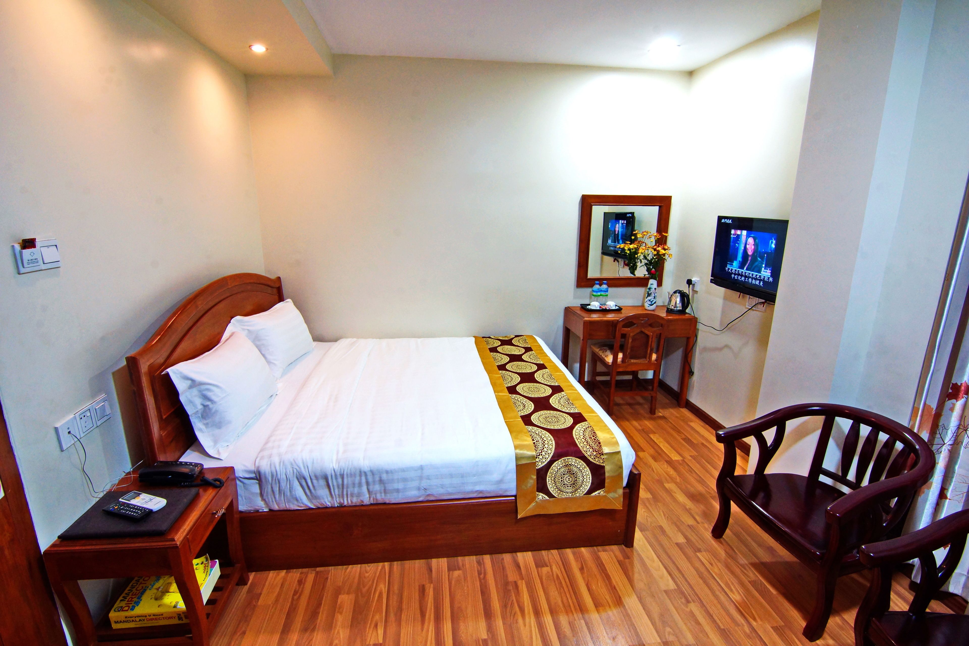 A hotel room with a wooden bed, a desk with a chair, a television, and a small table with a vase of flowers.