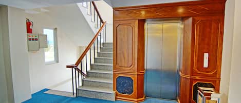 Una escalera con barandillas de madera, suelo cubierto de moqueta azul, un ascensor y un extintor de incendios montado en la pared.