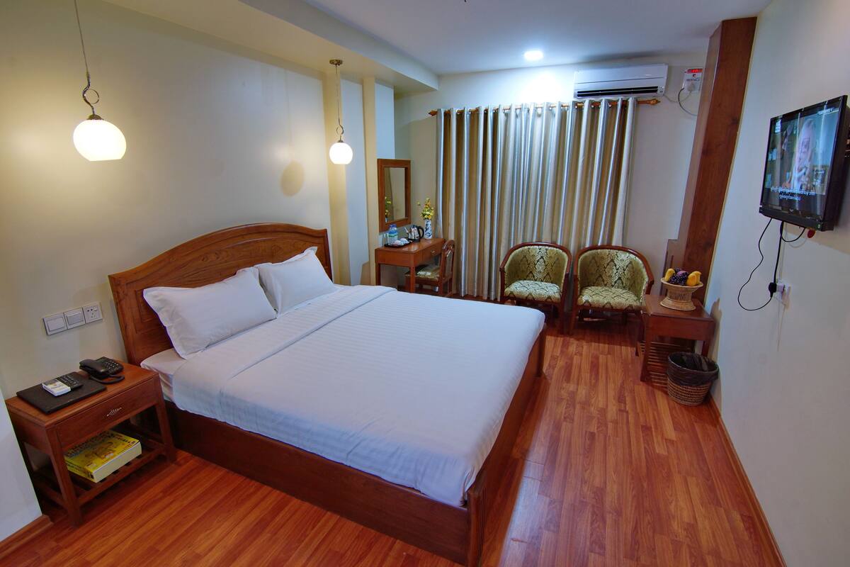 A neatly made bed with white linens, a wooden nightstand with a lamp, a desk with a chair, and a television mounted on the wall.