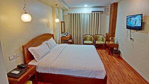 A hotel room with a large bed, bedside table with a phone and a book, a desk with a chair, a television mounted on the wall, and a curtain in the background. - Royal Pearl Hotel (Mandalay)