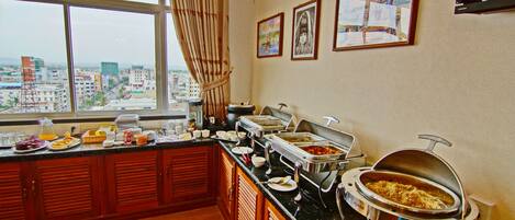 A buffet setup with various food stations, a large window with a view of the city, and artwork on the wall.