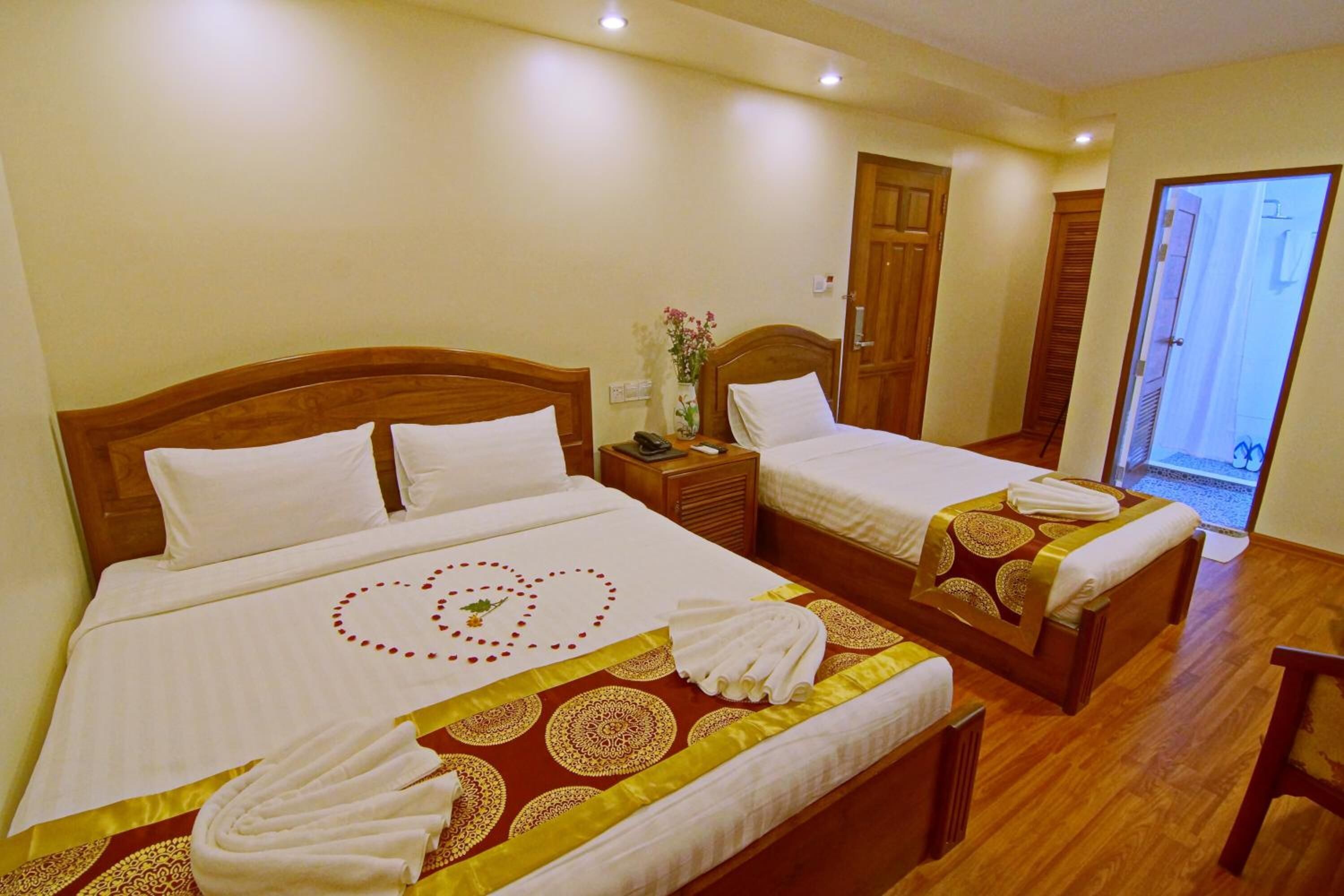 A hotel room with two beds, each with a wooden headboard, white bedding, and a decorative heart pattern on the bedspread. There is a wooden nightstand with a phone and a vase with flowers.