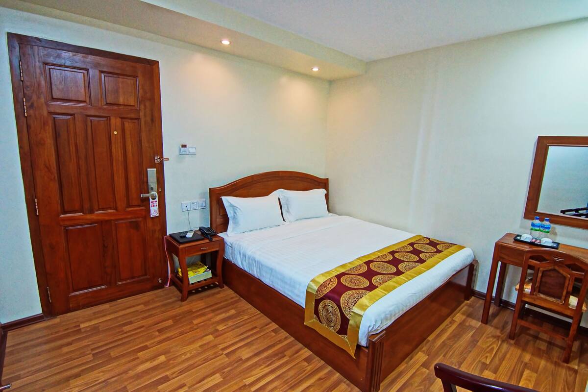 A hotel room with a wooden bed, bedside table, and a desk with a chair.