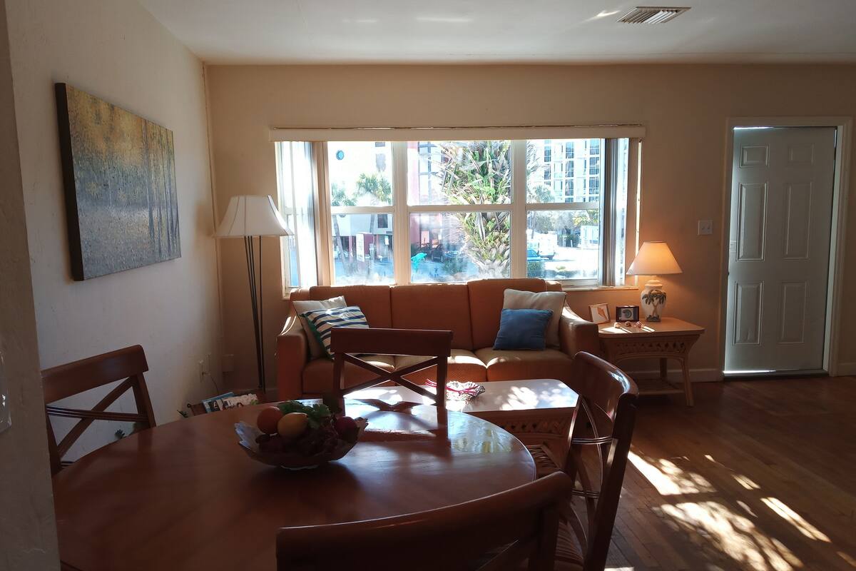 A dining area with a wooden table and chairs, a sofa with cushions, a lamp, and a window with a view of buildings and trees.