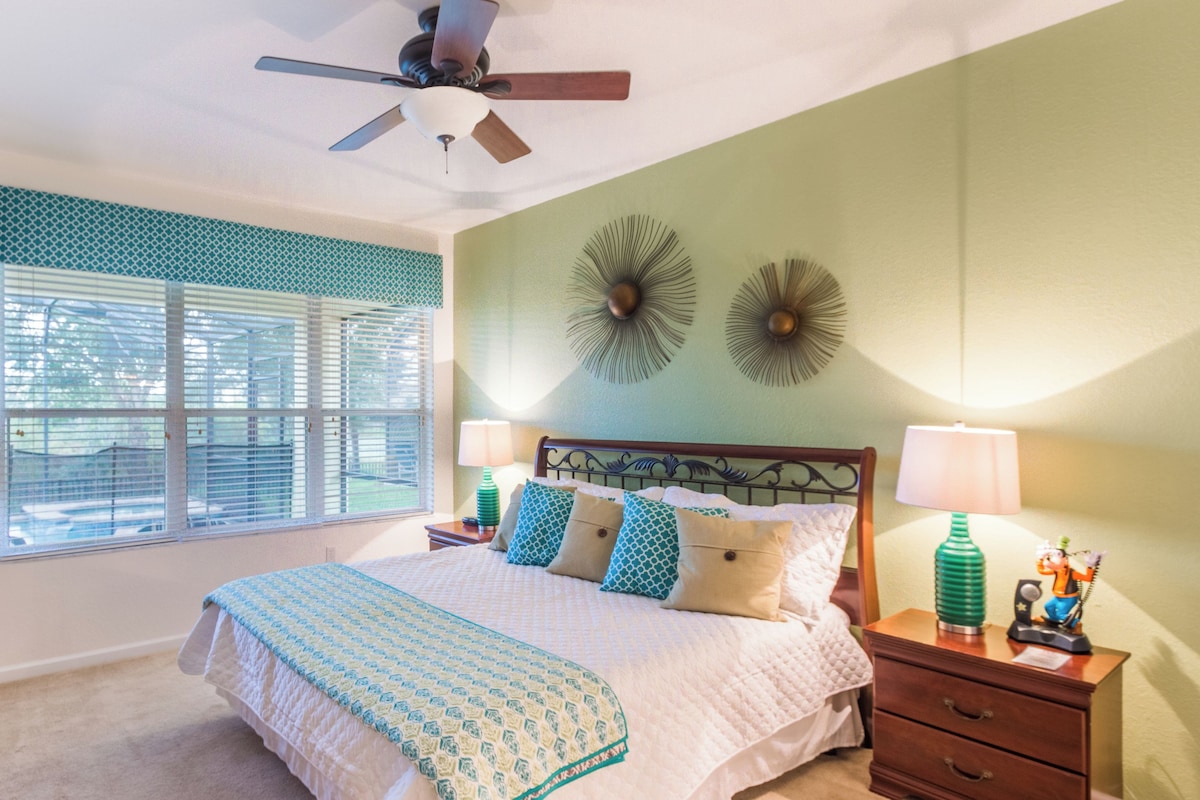 Une chambre à coucher comprenant un lit, des tables de chevet, un ventilateur de plafond, une fenêtre avec des stores et des éléments de décoration murale.