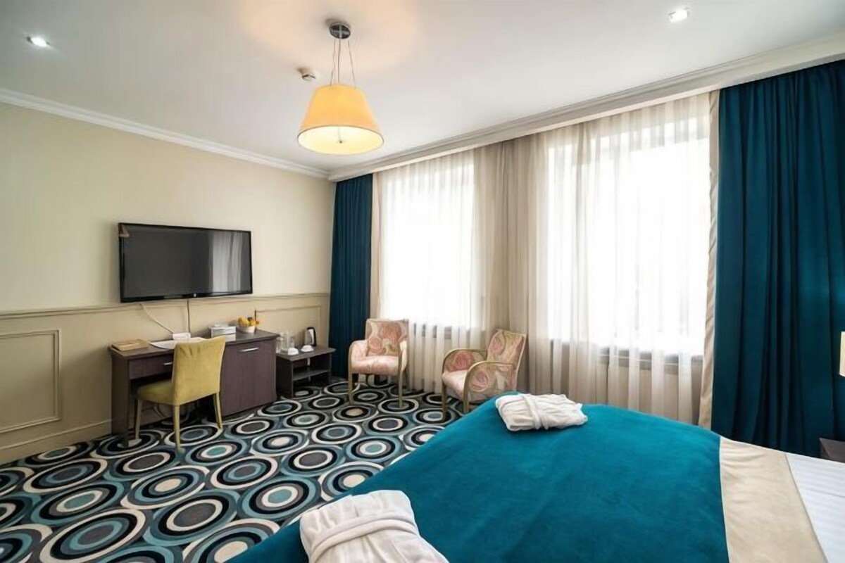 Habitación de hotel con cama, escritorio, silla, televisor y un amplio ventanal con cortinas.