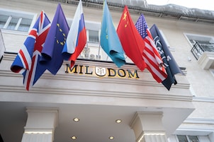El exterior de un hotel con varias banderas, incluyendo las de Reino Unido, la Unión Europea y Estados Unidos, y el nombre 'MILDOM' exhibido sobre la entrada.