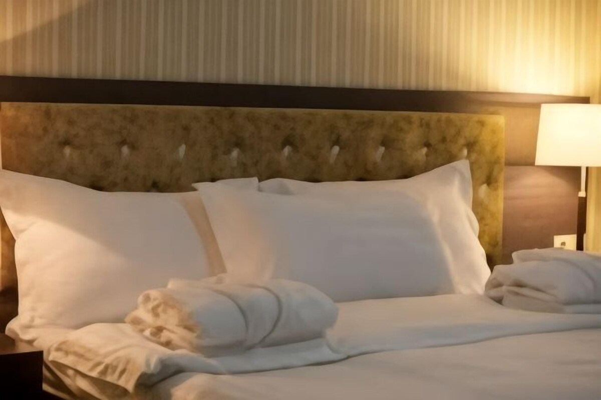 Una habitación de hotel con un cabecero capitonado, camas ordenadas con ropa blanca y una lámpara de noche.
