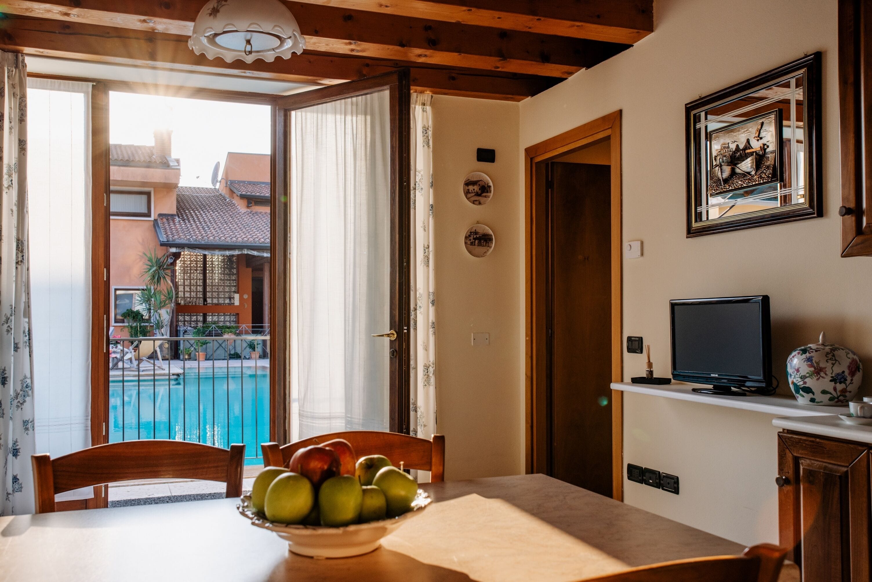 A dining area with a table, chairs, a bowl of fruit, a television, and a view of a swimming pool and buildings outside.