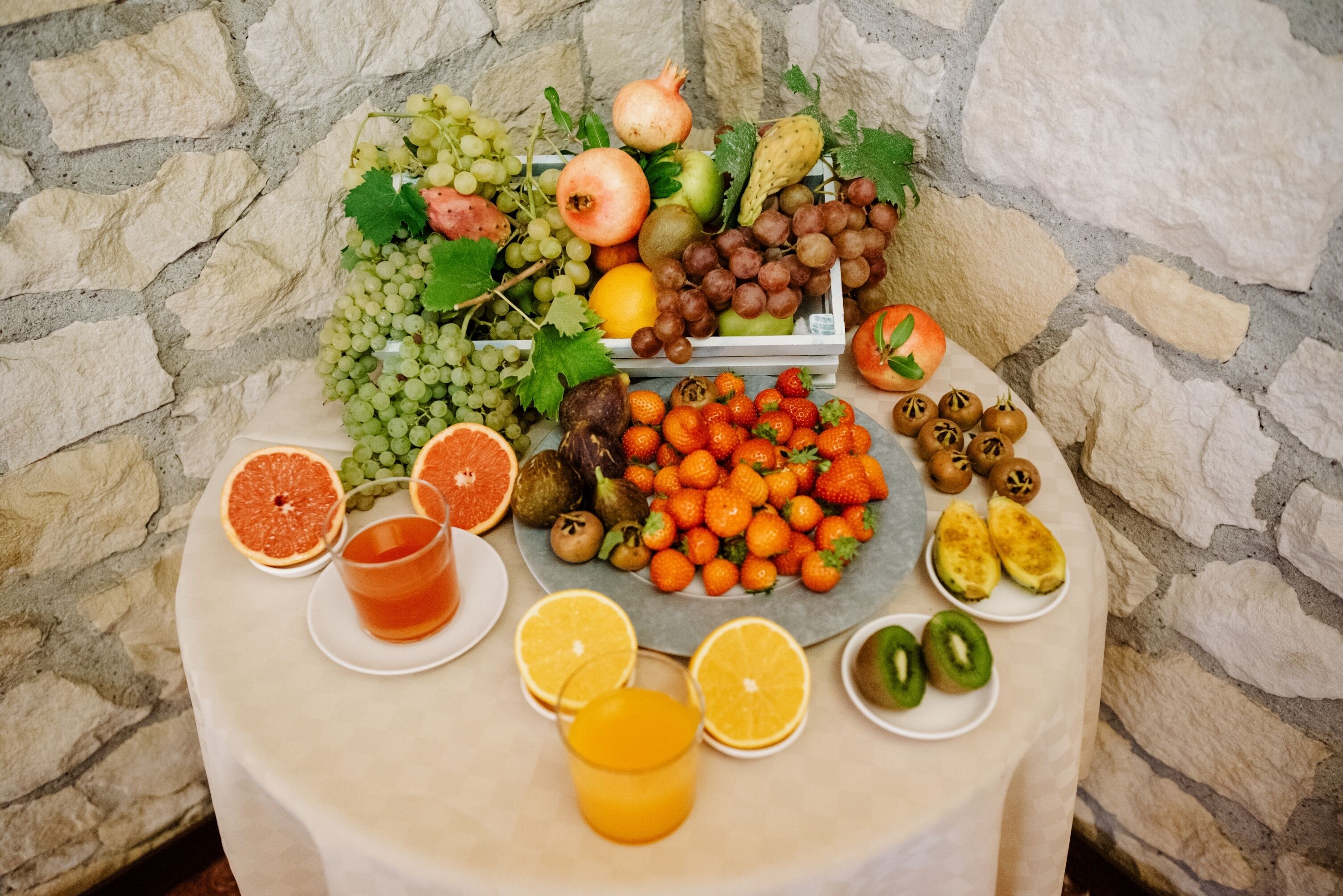 A table with a variety of fruits and beverages, including grapes, strawberries, and oranges, set against a stone wall background.