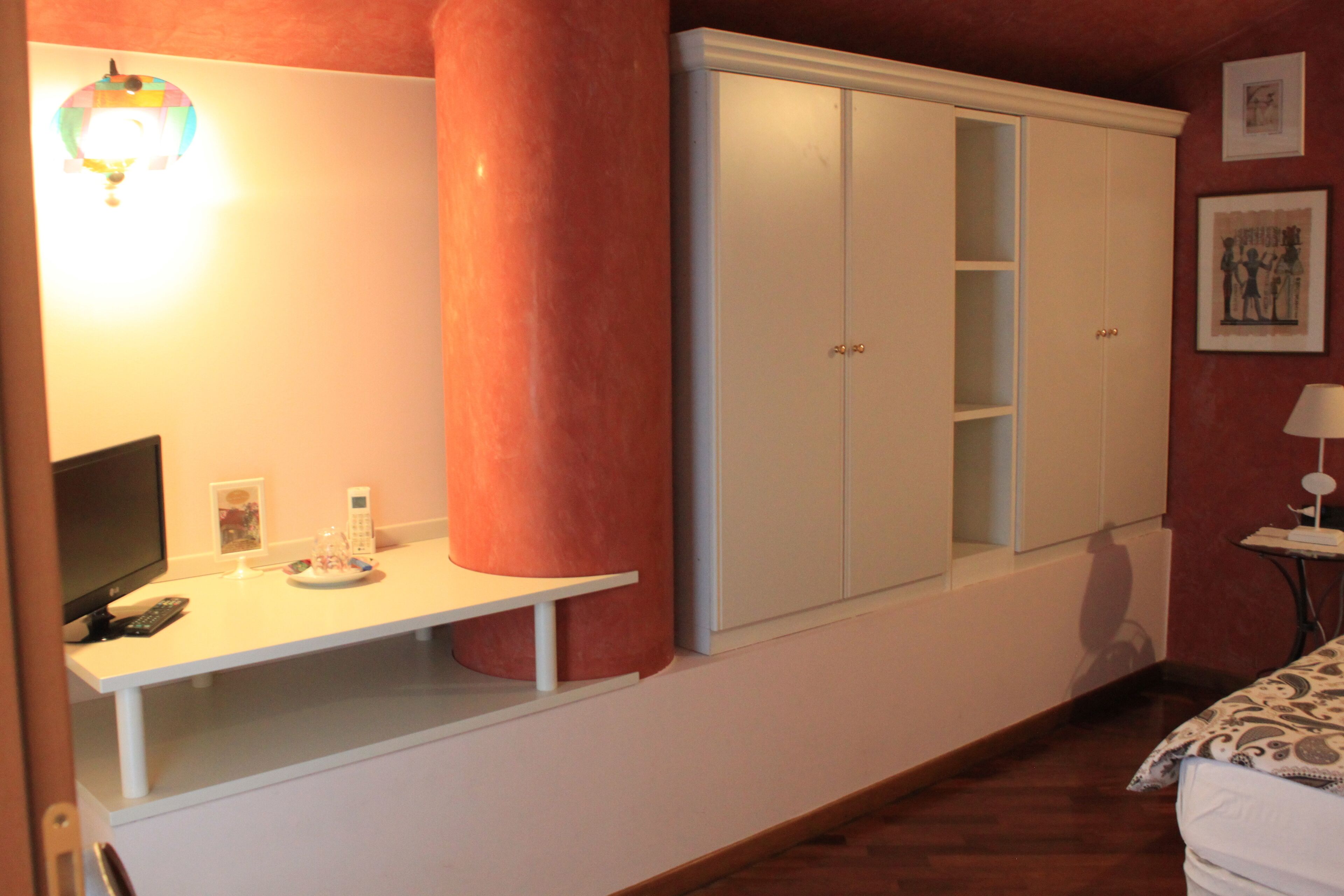 A bedroom with a bed, a desk with a lamp, a wardrobe, and a wall-mounted light fixture.