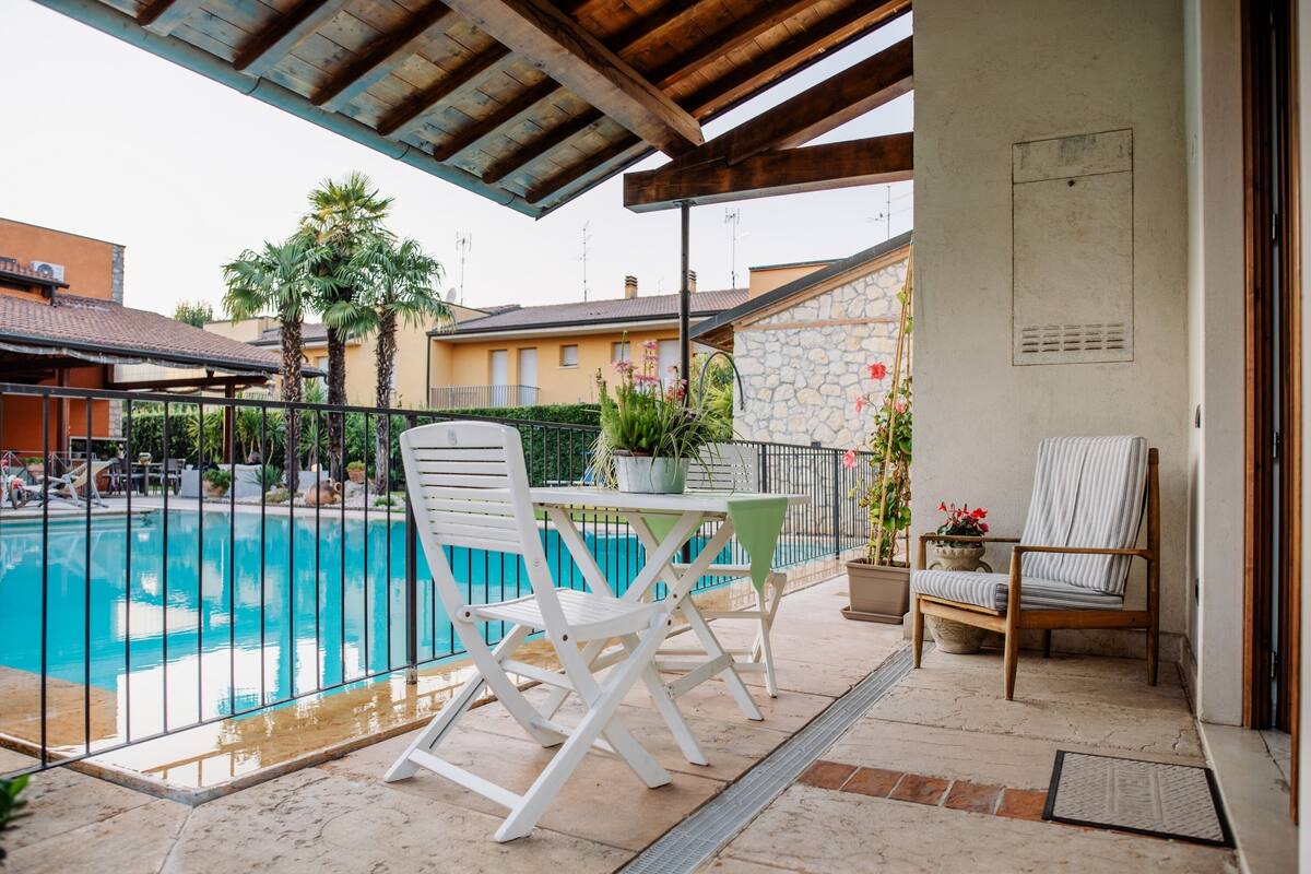 Un espace aménagé au bord de la piscine, avec un patio couvert, deux chaises blanches, une table et une plante en pot.