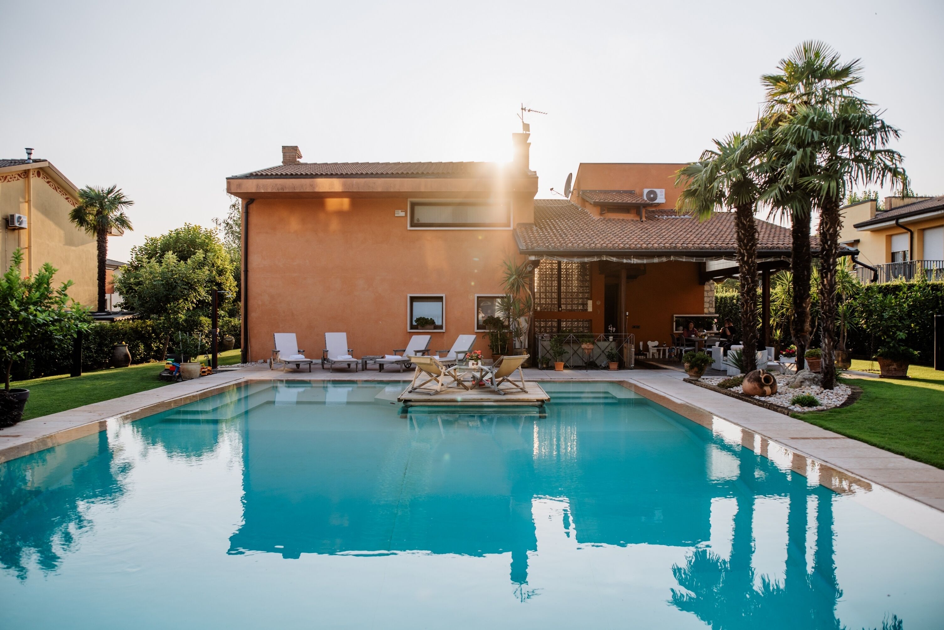 Una piscina con sillas de descanso y zona de patio, rodeada de palmeras y un edificio con tejado de baldosas.