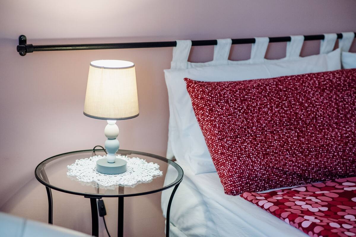 Un lit bien fait, avec des oreillers blancs et rouges, une table ronde en verre surmontée d'une nappe blanche et agrémentée d'une lampe, et une tête de lit en métal noir.