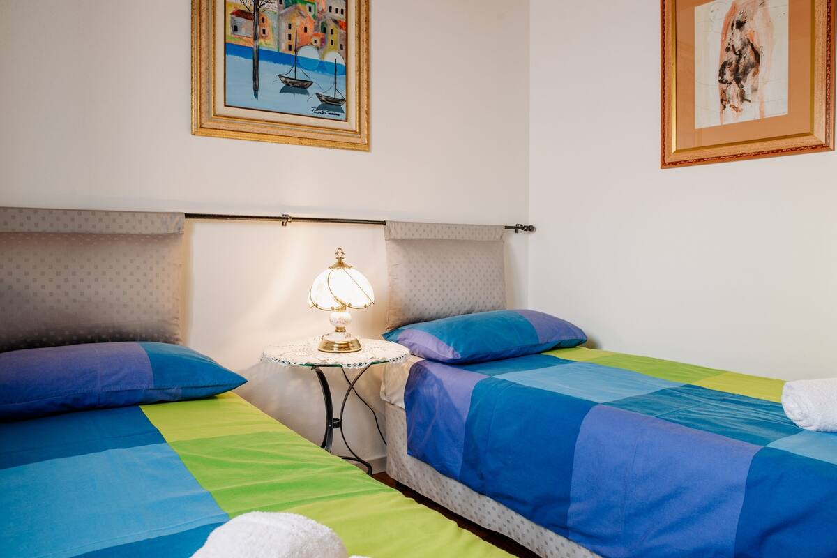 Une chambre d’hôtel avec deux lits, chacun agrémenté de draps bleus et verts, une petite table avec une lampe et deux tableaux accrochés aux murs.