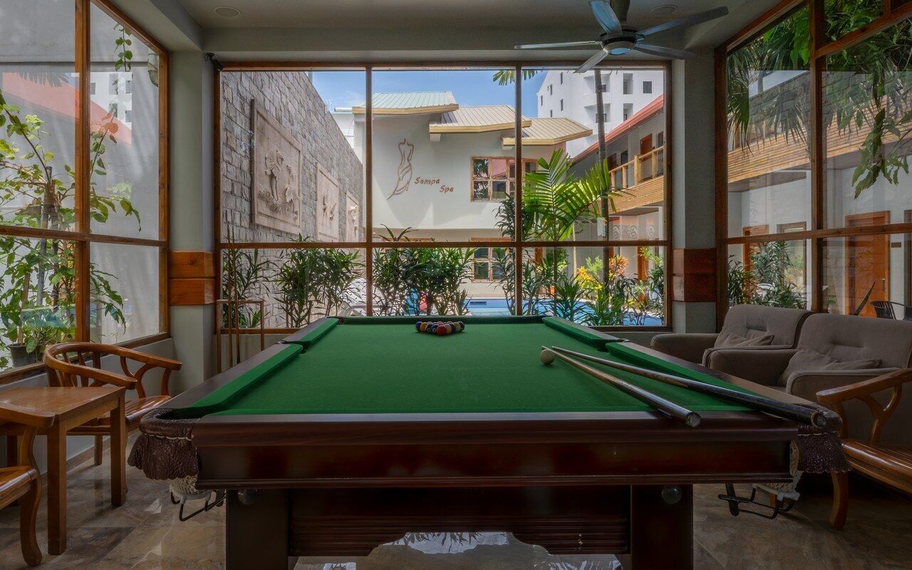 A pool table with surrounding wooden chairs, large windows, and indoor plants.