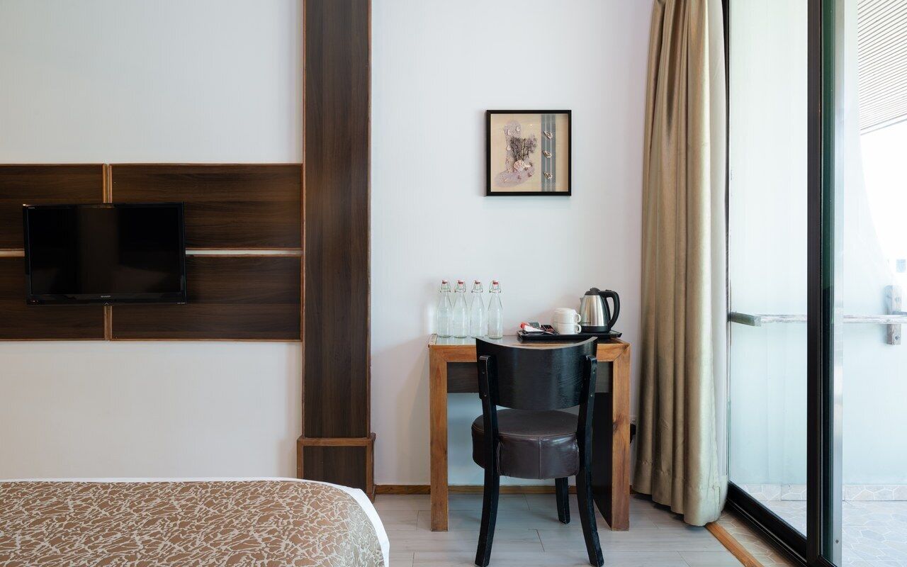 Habitación de hotel con cama, televisor montado en la pared, escritorio con silla, ventana con cortinas e imagen en un marco en la pared.