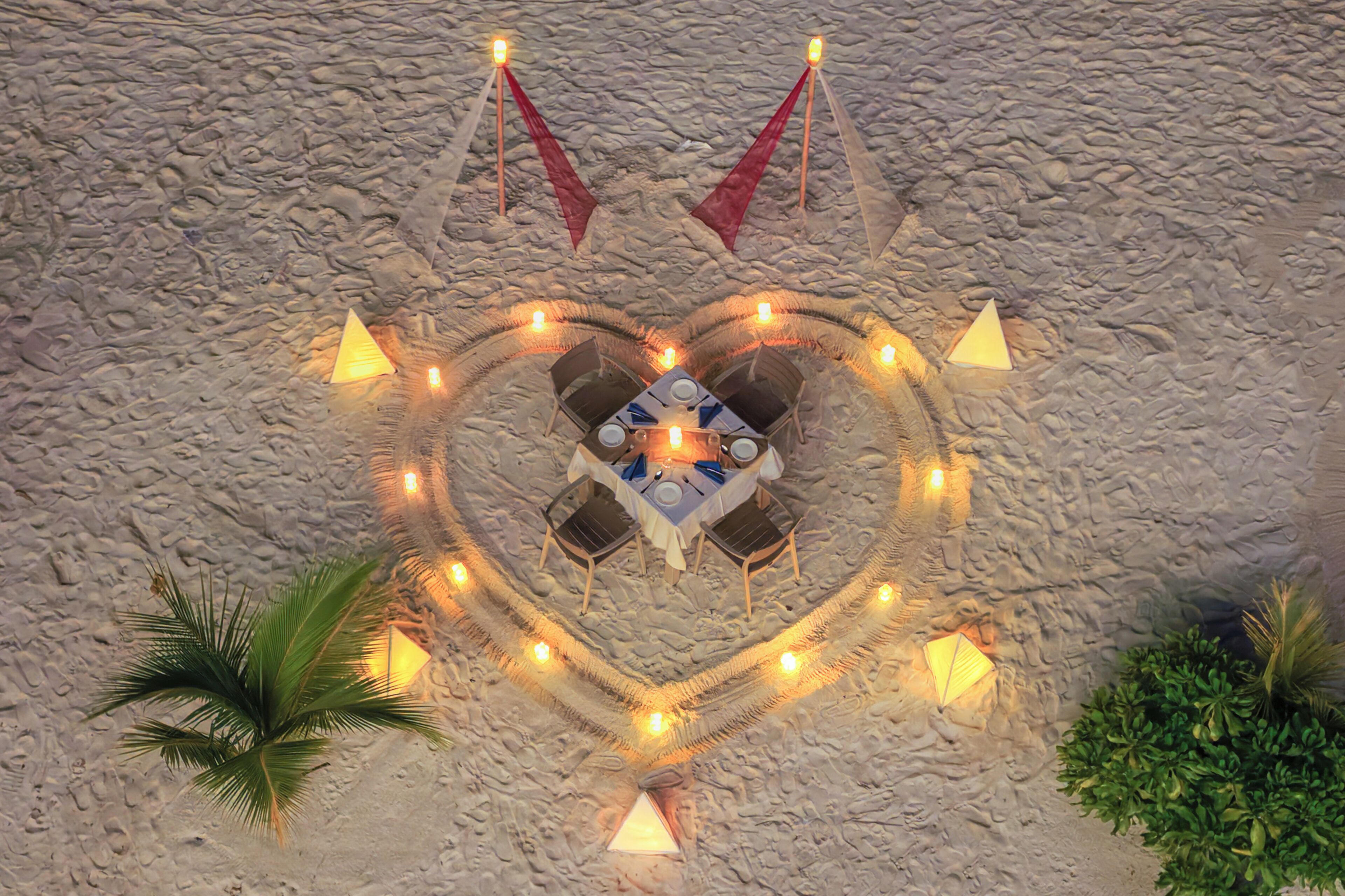 A heart-shaped arrangement of tables and chairs on a sandy surface, surrounded by string lights and candles.
