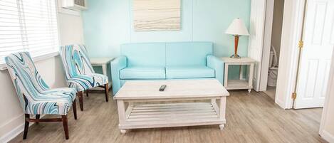 Uma sala de estar com um sofá azul claro, duas poltronas listradas, uma mesa de centro branca e um abajur sobre uma mesinha lateral.
