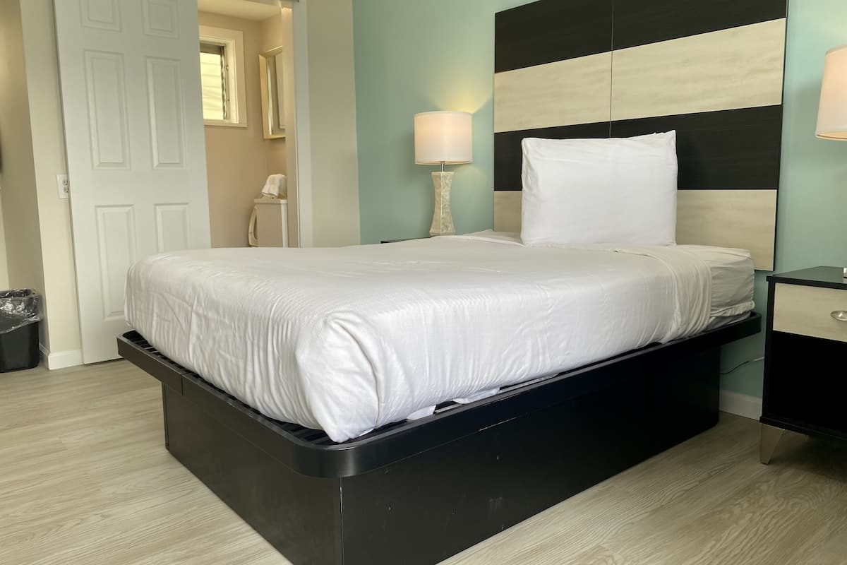 A neatly made bed with white linens in a room with a black and white headboard, a bedside lamp, and a trash bin.