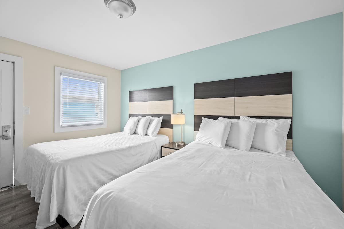 A hotel room with two beds, a nightstand, a lamp, and a window with blinds.