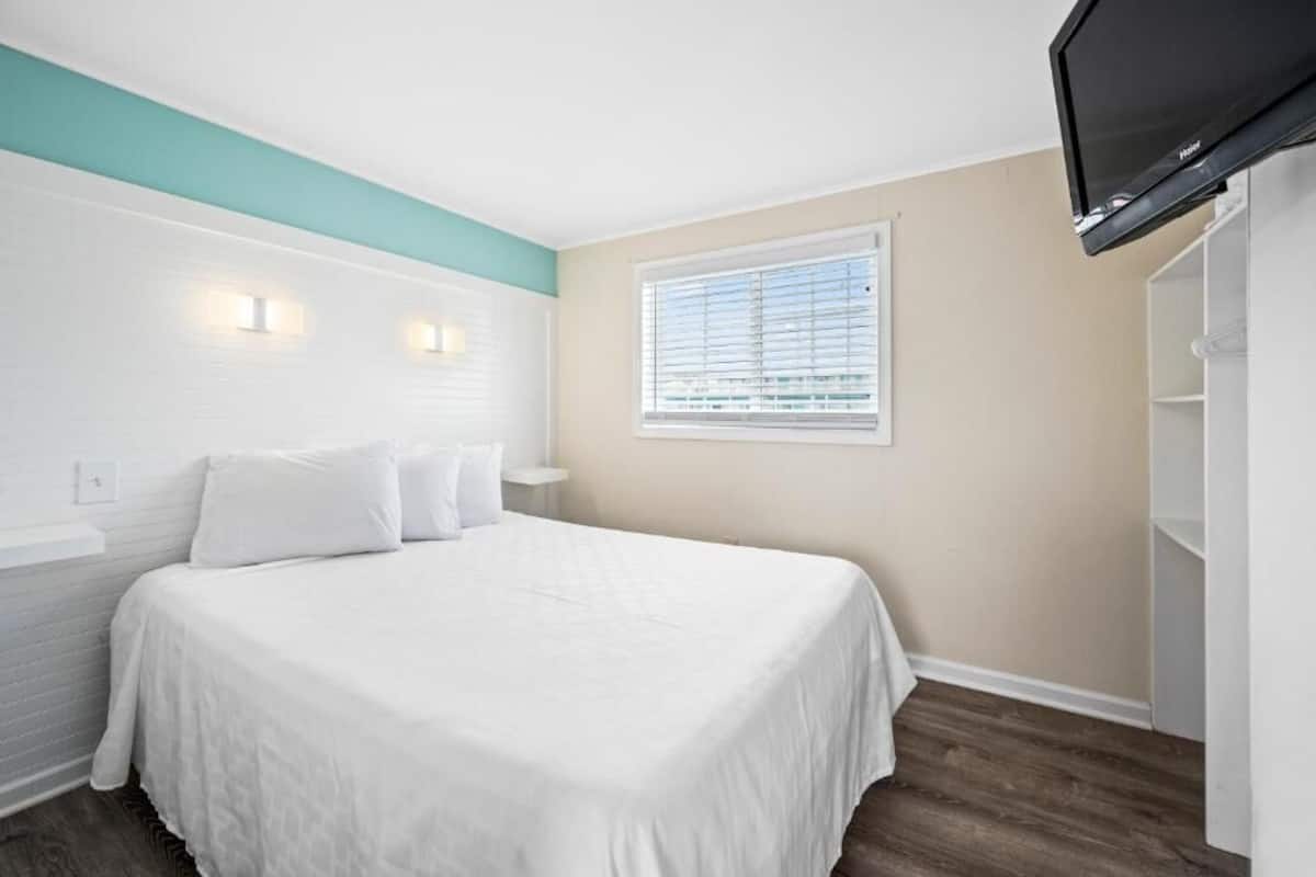 A bedroom with a bed, a window with blinds, a TV mounted on the wall, and a white wardrobe.