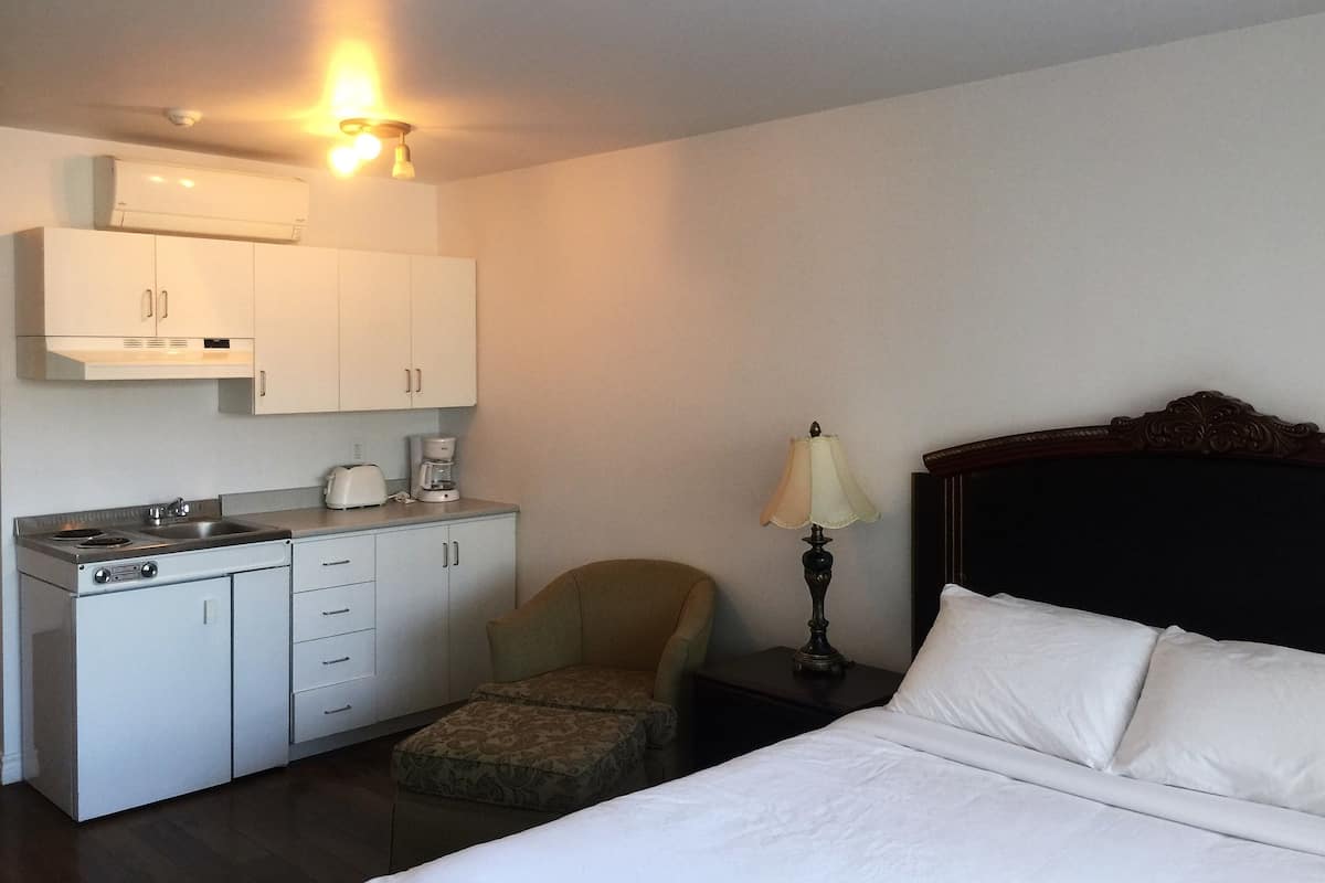 A hotel room with a bed, a small kitchenette with a stove, a chair, and a nightstand with a lamp.